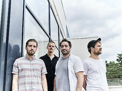 Bandfoto, die vier Musiker stehen vor einen gläsernen Gebäude