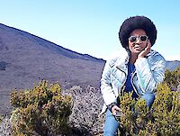 Eine Person sitzt in den Bergen. Sie trägt eine glänzende Jacke und eine Sonnenbrille. Um sie herum sind kleine Büsche und gelb-grüne Pflanzen. Im Hintergrund sieht man einen steilen Berghang und einen blauen Himmel. Die Person sieht entspannt aus.