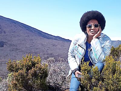 The picture shows a person wearing sunglasses and an iridescent jacket, sitting amidst low bushes in front of a barren mountain landscape. In the background, a steep mountainside can be seen under a blue sky, while the person strikes a relaxed pose and is surrounded by yellow-green vegetation.