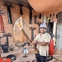 Ein Raum mit Ziegelwand zeigt viele schwarze Dinge. Es hängen Holz-Skulpturen und Handwerks-Sachen an der Wand. Eine Person steht im Vordergrund. Sie trägt ein beiges Hemd und einen bunten Hut. Die Person hält ein großes Holz-Musikinstrument wie eine Flöte.