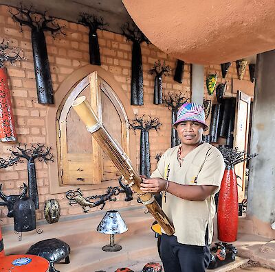 The picture shows a traditional craft room with a brick wall on which numerous black sculptural tree forms and handicraft objects hang. In the foreground, a person in a beige shirt and a colourful hat can be seen presenting a large, flute-like musical instrument made of wood.