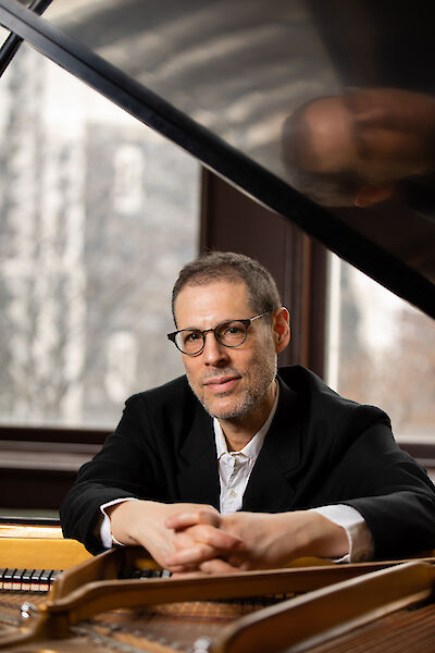 The picture shows a pianist in formal dress with glasses, sitting at a grand piano and resting on it with folded hands. A window with a view of buildings can be seen in the background, while the polished surface of the grand piano reflects the light.
