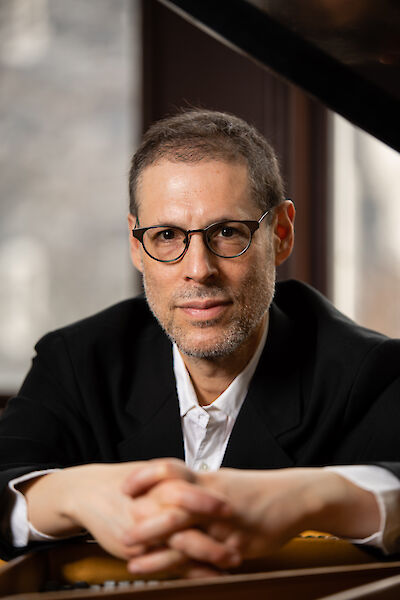 The picture shows a person with glasses in a black suit and white shirt sitting at a piano. The professional portrait is held in warm tones and presumably shows a pianist in a concentrated pose, his hands resting folded on the instrument.