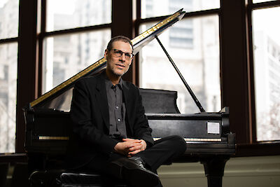 The picture shows a pianist in a black suit sitting in front of an open grand piano. Large windows can be seen in the background, providing a view of buildings in a city and allowing natural light into the room.
