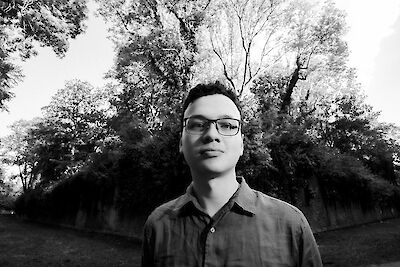 Das Bild zeigt eine Person. Die Person trägt ein Hemd und eine Brille. Im Hintergrund sind Bäume. Das Foto ist in Schwarz-Weiß. Die Aufnahme ist von unten gemacht. Es sieht ruhig aus.