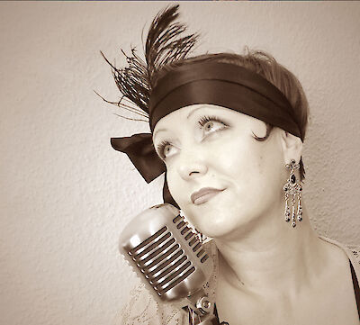The shot shows a person in a 1920s vintage style with a dark headband adorned with a feather, elegant earrings and stylish make-up next to a classic retro microphone. The sepia-coloured photo conveys a nostalgic jazz-era atmosphere and suggests a singer or performer in the style of the Roaring Twenties.
