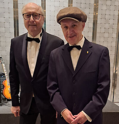 The picture shows two older gentlemen in elegant black suits with bow ties standing next to each other. The gentleman on the right is wearing a brown peaked cap and a small pin, while an electric guitar and a white lattice wall can be seen in the background.
