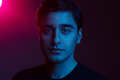 This image shows a portrait with dramatic lighting. The subject is lit by blue and pink light, creating a contrasting, modern atmosphere, while looking directly into the camera and wearing dark clothing.