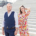 The picture shows a couple posing on a wide stone staircase. One person is wearing a blue waistcoat with a white shirt, while the other is wearing a colourful summer dress with a pink and yellow pattern and is holding a relaxed hand in her hair.