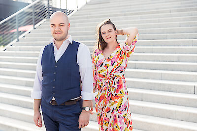 The picture shows a couple posing on a wide stone staircase. One person is wearing a blue waistcoat with a white shirt, while the other is wearing a colourful summer dress with a pink and yellow pattern and is holding a relaxed hand in her hair.