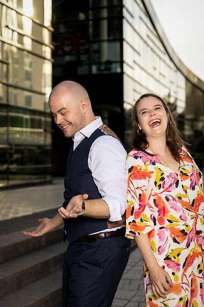 The picture shows two people laughing happily in front of a modern building with a glass façade. The person on the left is wearing a dark blue waistcoat over a white shirt, while the person on the right is wearing a colourful dress with a floral pattern in orange, pink and yellow and gold earrings.