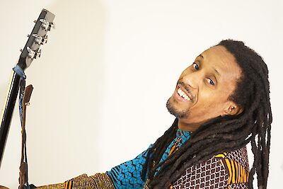 Das Bild zeigt einen Musiker mit Dreadlocks in einem farbenfrohen, gemusterten Outfit, der lächelt während neben ihm der Hals einer Gitarre zu sehen ist. Der Hintergrund ist hell und schlicht gehalten, was den Fokus auf den Musiker und sein Instrument richtet.