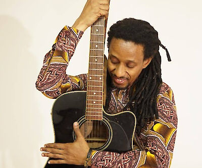 Das Bild zeigt eine Person mit Dreadlocks, die eine schwarze Akustikgitarre spielt und dabei ein farbenprächtiges Hemd mit rot-goldenen Mustern trägt. Die Person hält die Gitarre in einer emotionalen, intimen Pose und scheint in die Musik vertieft zu sein, während sie vor einem hellen Hintergrund fotografiert wurde.