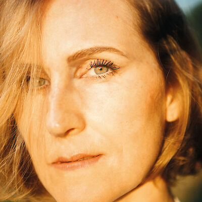 Das Bild zeigt ein nahes Portrait mit einem Fokus auf Augen und Gesichtszüge in warmem Sonnenlicht. Die blonde Haarpracht und die natürlich wirkende Haut werden von einem goldenen Licht umspielt, das dem Bild eine atmosphärische Wärme verleiht.