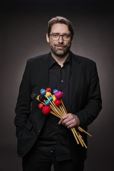 Das Bild zeigt eine Person in einem schwarzen Anzug mit schwarzem Hemd, die eine Sammlung von Mallets oder Schlägeln für ein Percussion-Instrument hält. Die Schlägel haben bunte Köpfe in verschiedenen Farben wie Rot, Blau, Gelb und Pink, die an hölzernen Stielen befestigt sind, vor einem neutral grauen Hintergrund.