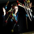 Ein Mann steht an einem Instrument. Das Instrument heißt Vibraphon. Der Mann trägt ein blaues Hemd und ein dunkles Jackett. Um ihn herum sind Schlägel. Die Schlägel haben rote, blaue und gelbe Köpfe. Die Schlägel bewegen sich schnell. Der Mann spielt konzentriert. Die Bewegung der Schlägel zeigt die Energie der Musik.