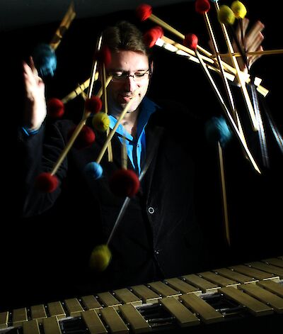 Das Bild zeigt einen Musiker in einem dunklen Raum, der an einem Vibraphon oder Marimbaphon steht, während Schlägel mit roten, blauen und gelben Köpfen um ihn herum in Bewegung erscheinen. Die Person trägt ein blaues Hemd unter einem dunklen Sakko und ist konzentriert auf das Instrument vor sich gerichtet, während eine dynamische Bewegungsunschärfe der Schlägel die Energie des musikalischen Moments einfängt.