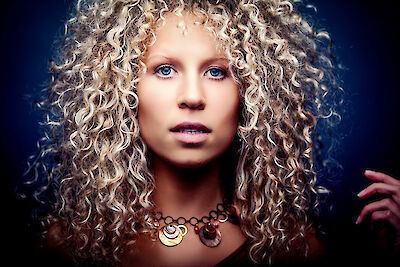The image shows a person with voluminous, spiralling blonde curls against a dark blue background. The person is wearing an eye-catching necklace with round pendants and the shot shows intense blue eyes and an artistic portrait with dramatic lighting.