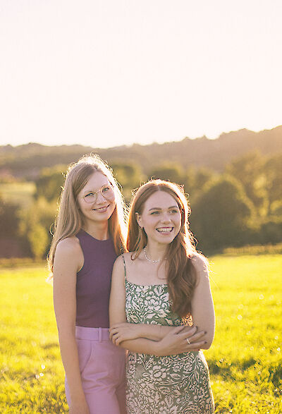 Das Bild zeigt zwei Personen in einem sonnendurchfluteten Feld während des goldenen Sonnenuntergangslichts. Eine Person trägt ein lila Oberteil mit rosa Hose, während die andere ein geblümtes Kleid trägt, beide stehen vor einer malerischen Landschaft mit sanften Hügeln und Bäumen im Hintergrund.