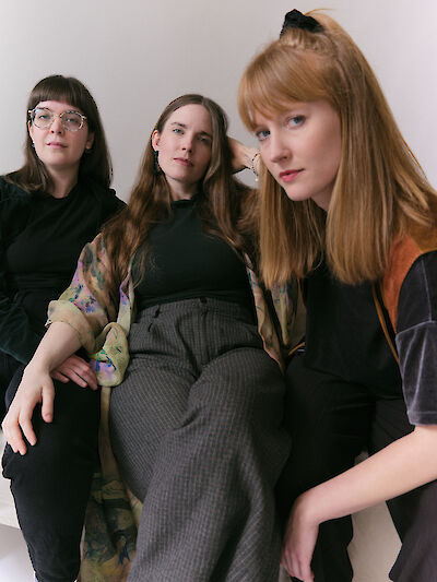 The picture shows three people with long hair in stylish, predominantly dark-coloured clothing against a light background. The person on the left is wearing round glasses, the person in the middle has a patterned top over a black top, while the person on the right has an orange accent in their clothing.