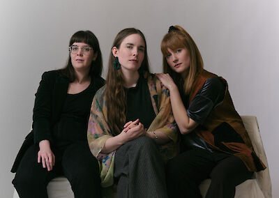 The picture shows three people in elegant dark clothing posing on a light-coloured seat in front of a plain white wall. The person in the middle is wearing a black top with a colourful scarf, while the person on the left is wearing glasses and the person on the right is wearing a combination of black clothing with colourful accents.