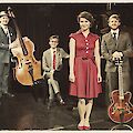 Das Bild zeigt Musiker. Sie spielen Jazz. Die Musiker tragen alte Kleider. Links steht ein großer Musik-Bogen. In der Mitte ist ein Klavier. Eine Frau trägt ein rotes Kleid. Rechts ist eine Gitarre. Alle Musiker sehen schick aus.