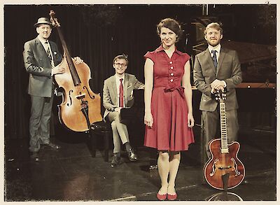 Das Bild zeigt ein Jazzensemble in Vintage-Stil-Kleidung auf einer Bühne mit verschiedenen Instrumenten. Zu sehen sind ein Kontrabass links, ein Piano in der Mitte, eine Person in einem roten Kleid im Vordergrund und eine Jazzgitarre rechts, wobei alle Musiker in eleganter Kleidung posieren.