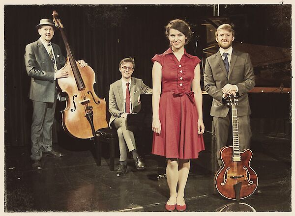 The picture shows a jazz ensemble in vintage-style clothing on a stage with various instruments. You can see a double bass on the left, a piano in the centre, a person in a red dress in the foreground and a jazz guitar on the right, with all the musicians posing in elegant clothing.