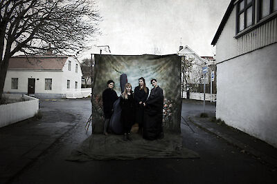 Das Bild zeigt ein Kunst-Foto. Es ist auf einer Straße zwischen weißen Häusern. 4 Personen tragen schwarze Kleidung. Sie stehen vor einem dunklen Hintergrund. Das Bild sieht aus wie eine kleine Bühne.