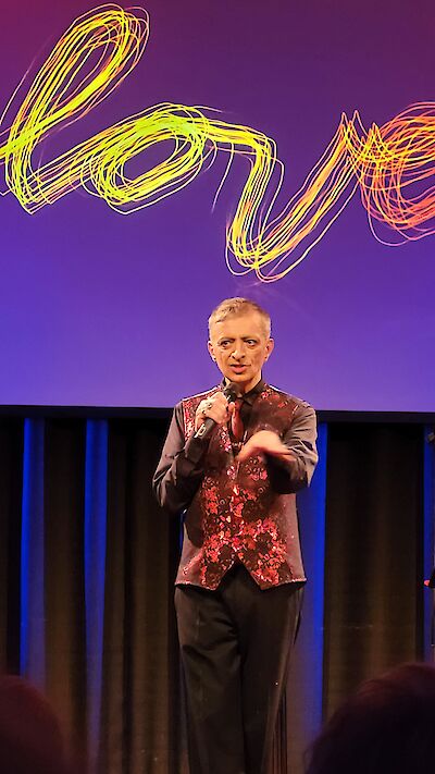 The image shows a person on a stage holding a microphone and wearing a striking red patterned waistcoat over a dark shirt. In the background is a projection screen with bright, yellow-orange "love" lettering in a dynamic, curved light font.