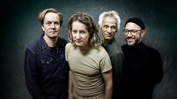 The picture shows four people standing in front of a dark grey, circular background. The group consists of two people with light-coloured hair in dark and light-coloured clothing, one person with white hair in olive-coloured clothing and one person with glasses and a cap in black clothing, who together look like a musical ensemble or a group of artists.