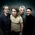 The picture shows four people standing in front of a dark, circular background and looking into the camera. The group consists of two people with short hair, one person with longer, wavy hair in a beige T-shirt in the centre, and an older person with white hair, all wearing different items of clothing such as a denim shirt, T-shirt and turtleneck jumper.
