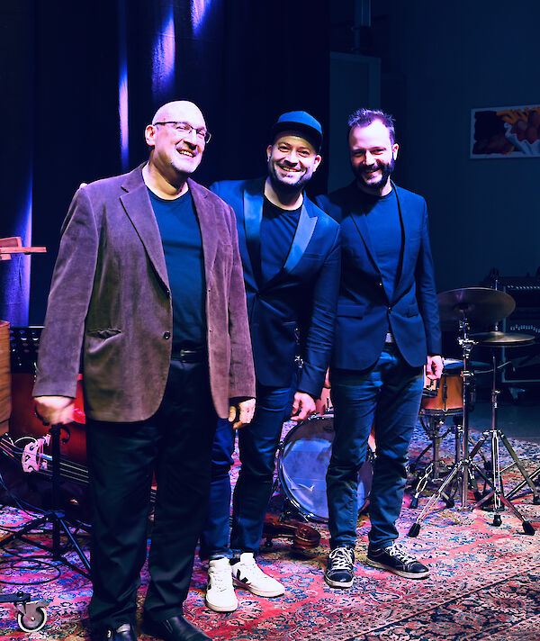 The image shows three people on a stage posing together, all dressed in dark blues and browns and smiling. Musical instruments can be seen in the background, including a drum kit and a guitar, suggesting that this is a musical event or concert.