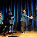 The picture shows a stage scene with four people in front of a blue curtain, with two people shaking hands, while one person sits at the keyboard and another stands in the background. The scene takes place on a wooden stage where an event such as a concert, theatre or award ceremony is presumably taking place.
