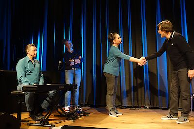 The picture shows a stage scene with four people in front of a blue curtain, with two people shaking hands, while one person sits at the keyboard and another stands in the background. The scene takes place on a wooden stage where an event such as a concert, theatre or award ceremony is presumably taking place.