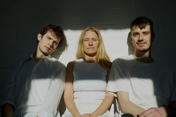 The picture shows three people in light-coloured clothing standing in front of a white wall, with interesting light and shadow effects. The group stands next to each other in an artistic pose, with the person in the centre wearing a white outfit and flanked by two other people in light-coloured shirts.