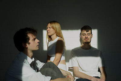 Das Bild zeigt drei Personen in einem minimalistischen, von Licht und Schatten geprägten Raum, wobei links eine Person in einem weißen Hemd sitzt, in der Mitte eine Person in weißer Kleidung steht und rechts eine weitere Person in einem weißen T-Shirt mit geschlossenen Augen sitzt. Die dramatische Beleuchtung erzeugt einen künstlerischen Kontrast zwischen den hellen Kleidungsstücken und dem dunklen Hintergrund, wobei ein Lichtstrahl von rechts einen Teil der Szene erhellt.