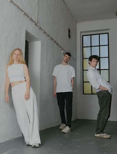 Das Bild zeigt drei Personen in einem minimalistischen, weiß gestrichenen Raum mit einem großen Fenster. Alle tragen überwiegend weiße Kleidung - die Person links in einem weißen Crop-Top und weiter Hose, die mittlere Person in einem weißen T-Shirt mit dunkler Hose und die rechte Person in einem weißen Hemd mit grüner Jeans.