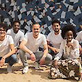 The picture shows a group of six people posing in a row in front of a mosaic-like wall with blue, grey and orange tiles. The people are predominantly wearing white T-shirts and light-coloured trousers, with one person in the centre right wearing a colourful, flowered skirt, while they all smile at the camera and squat or kneel on the sandy floor.