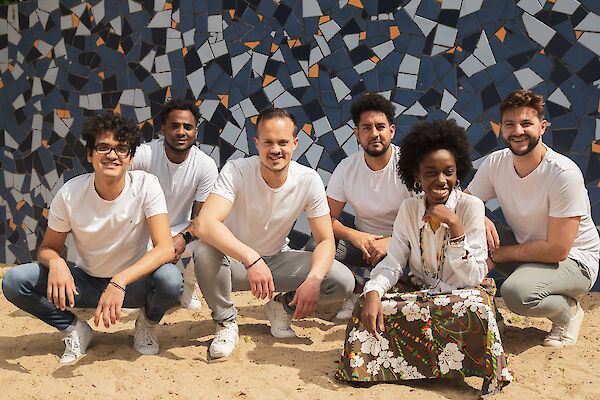The picture shows a group of six people posing in a row in front of a mosaic-like wall with blue, grey and orange tiles. The people are predominantly wearing white T-shirts and light-coloured trousers, with one person in the centre right wearing a colourful, flowered skirt, while they all smile at the camera and squat or kneel on the sandy floor.