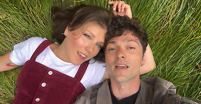 Das Bild zeigt zwei Personen, die in einem hohen Grasfeld liegen und ein Selfie machen. Eine Person trägt einen weißen Pullover mit einem burgunderroten Latzhosenkleid, während die andere Person ein graues Oberteil trägt, beide ruhen entspannt im grünen Gras.