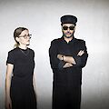 Das Bild zeigt zwei Personen in schwarzer Kleidung vor einer neutralen, grauen Wand. Die Person auf der linken Seite trägt eine Brille und ein schwarzes Outfit, während die Person auf der rechten Seite eine schwarze Kapitänsmütze, eine Sonnenbrille und ein schwarzes Hemd trägt und mit verschränkten Armen posiert.
