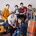 Das Bild zeigt eine Gruppe junger Menschen in einem künstlerischen Setting mit verschiedenen Musikinstrumenten und Dekorationselementen. Im Vordergrund ist ein orangefarbenes Keyboard zu sehen, während im Hintergrund Pflanzen, eine kleine Leiter und andere kreative Objekte arrangiert sind, was auf eine Band oder ein Musikensemble hindeutet.