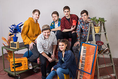 Das Bild zeigt eine Gruppe junger Menschen in einem künstlerischen Setting mit verschiedenen Musikinstrumenten und Dekorationselementen. Im Vordergrund ist ein orangefarbenes Keyboard zu sehen, während im Hintergrund Pflanzen, eine kleine Leiter und andere kreative Objekte arrangiert sind, was auf eine Band oder ein Musikensemble hindeutet.