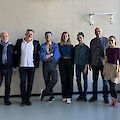 Das Bild zeigt eine Gruppe von sieben Personen, die nebeneinander vor einer grauen Betonwand posieren. Die Gruppe steht in einem Raum mit Ballettstange, vermutlich in einem Tanzstudio oder einer künstlerischen Einrichtung, und trägt verschiedene Kleidung in überwiegend blauen und dunklen Farbtönen.