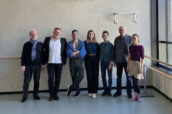 Das Bild zeigt eine Gruppe von sieben Personen, die nebeneinander vor einer grauen Betonwand posieren. Die Gruppe steht in einem Raum mit Ballettstange, vermutlich in einem Tanzstudio oder einer künstlerischen Einrichtung, und trägt verschiedene Kleidung in überwiegend blauen und dunklen Farbtönen.