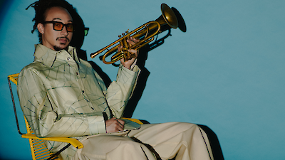 Das Bild zeigt einen Musiker in einem beigen Outfit, der auf einem gelben Stuhl sitzt und eine goldene Trompete in der Hand hält. Der Hintergrund ist in einem türkisblauen Farbton gehalten, was einen schönen Kontrast zu dem beigen Outfit und dem goldenen Instrument bildet.