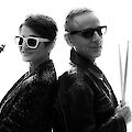 The picture shows two musicians posing back to back in an artistic black and white shot. The person on the left is wearing white sunglasses and holding a saxophone, while the person on the right is wearing dark sunglasses and holding drumsticks, both wearing dark clothing, suggesting a jazz or rock duo.