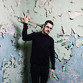 The picture shows a musician in black clothing holding a trumpet over his shoulder. He is standing in front of a heavily peeling wall of mint green and blue paint, which forms an interesting, weathered background.