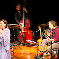 Das Bild zeigt eine Jazz-Band während eines Auftritts auf einer beleuchteten Bühne. Im Vordergrund sieht man eine Sängerin in einem lila Kleid mit Mikrofon, während im Hintergrund Musiker mit Kontrabass, Gitarre, Schlagzeug und Klavier zu sehen sind.